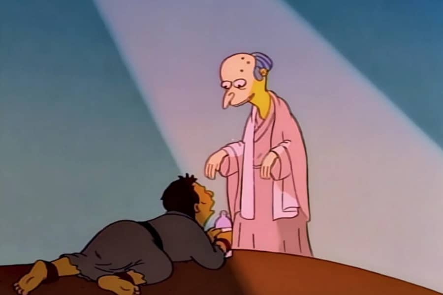 a man lays in the desert sand at the feet of Mr. Burns who is standing in a God-like beam of light