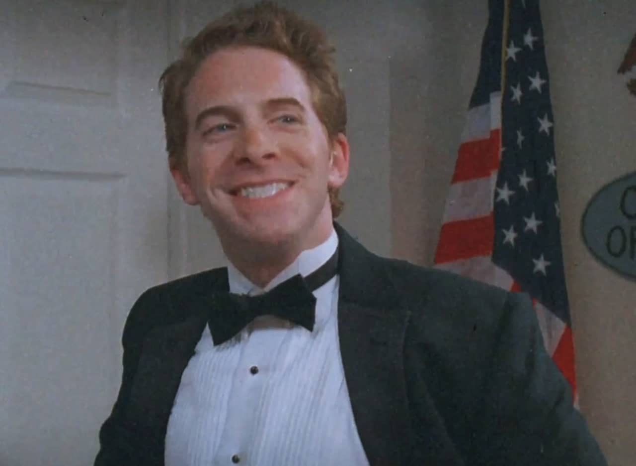 Buddy Zaks smiling in a tuxedo, American flag behind