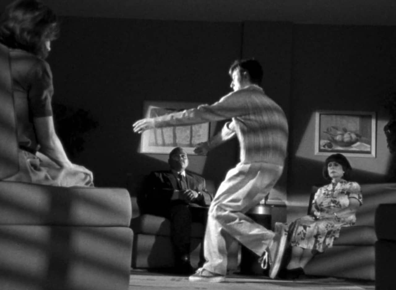 a black and white scene of a man dancing in a living room surrounded by a stoic family