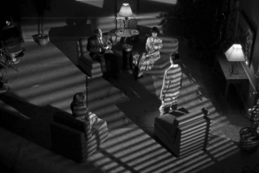 the man stands in front of the family, moonlight casting stripes across the room through window blinds