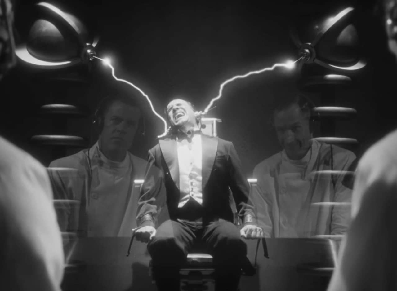 a man in a tuxedo is strapped to a chair and screams while two men in lab coats send electric jolts to his head