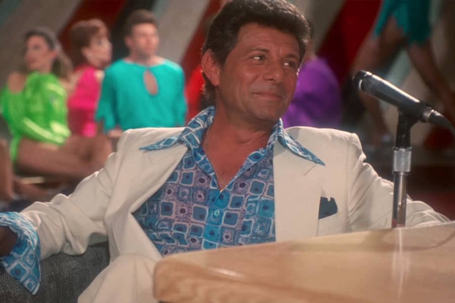 a guest in a patterened shirt with wide collar and white suit