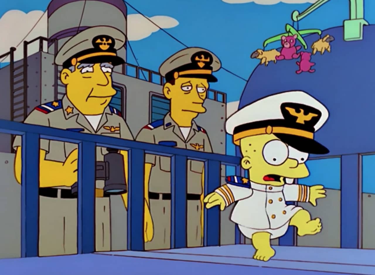 two Navy men on a ship next to a crib with a baby wearing an admiral’s uniform