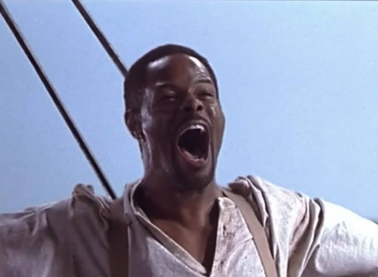 a Black man on a boat raises his arms and yells triumphantly