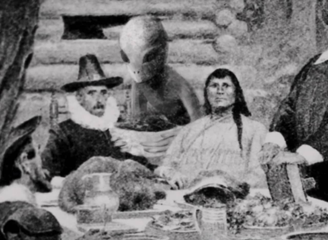 a grainy photograph of the Pilgrims eating Thanksgiving dinner with the Native Americans as an alien serves them food