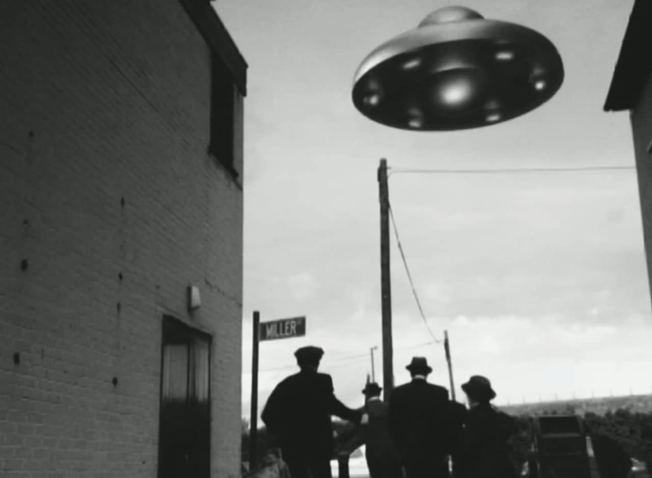 men run down an alley as an alien ship hovers in the sky