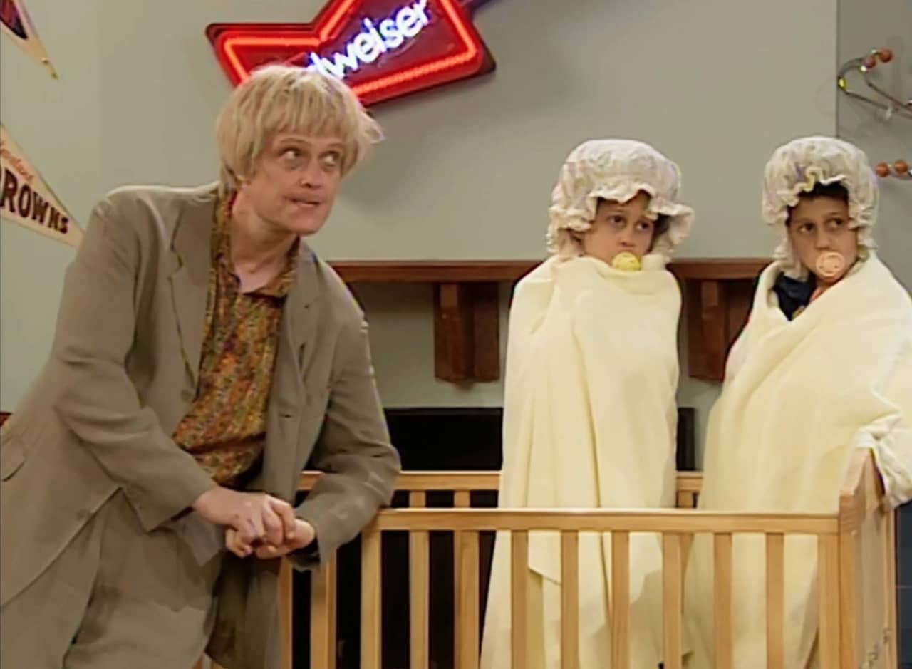 Dieter Dalmer in a tan suit and scraggly blonde hair, next to two non-toddler kids dressed as babies in a crib