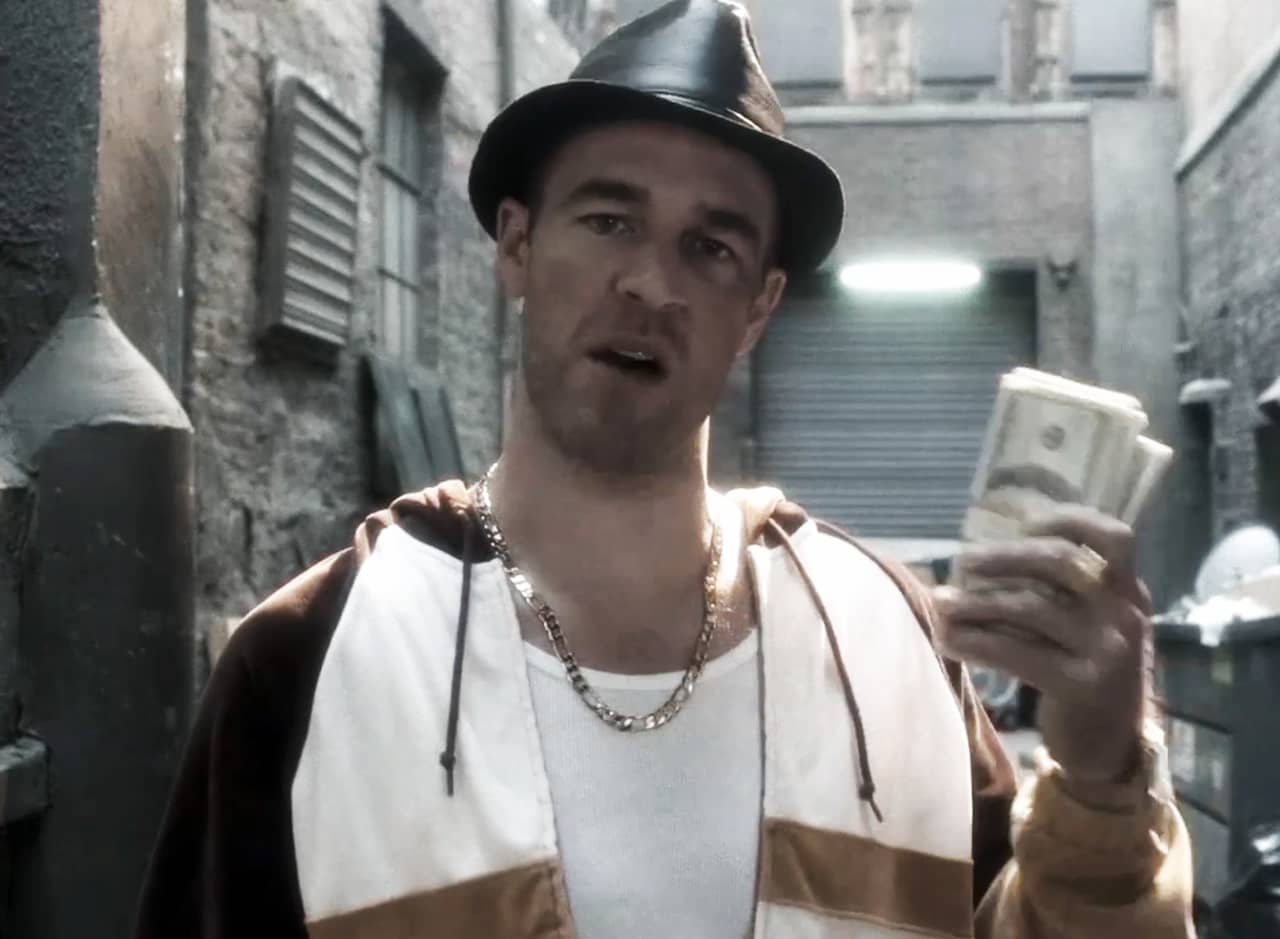 James Van Der Beek in a dirty alley, wearing a big chain and fedora, holding up a stack of cash