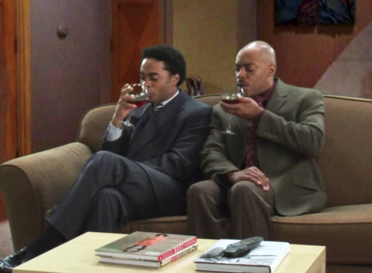 two Black men in suits sit on a couch enjoying glasses of red wine