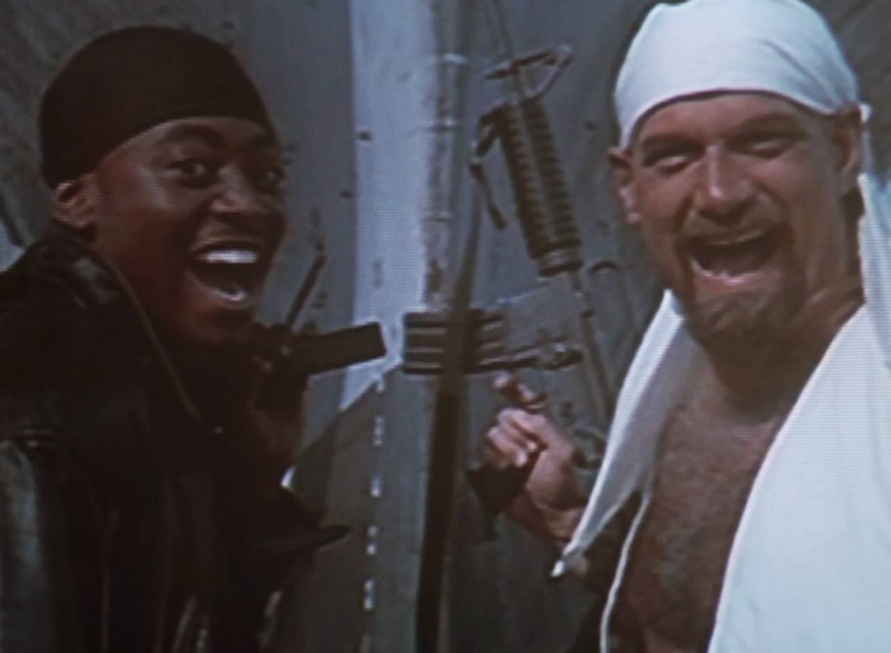 a Black man (Willie Mays Hays) and white man (Jesse Ventura) stand together smiling big while holding guns