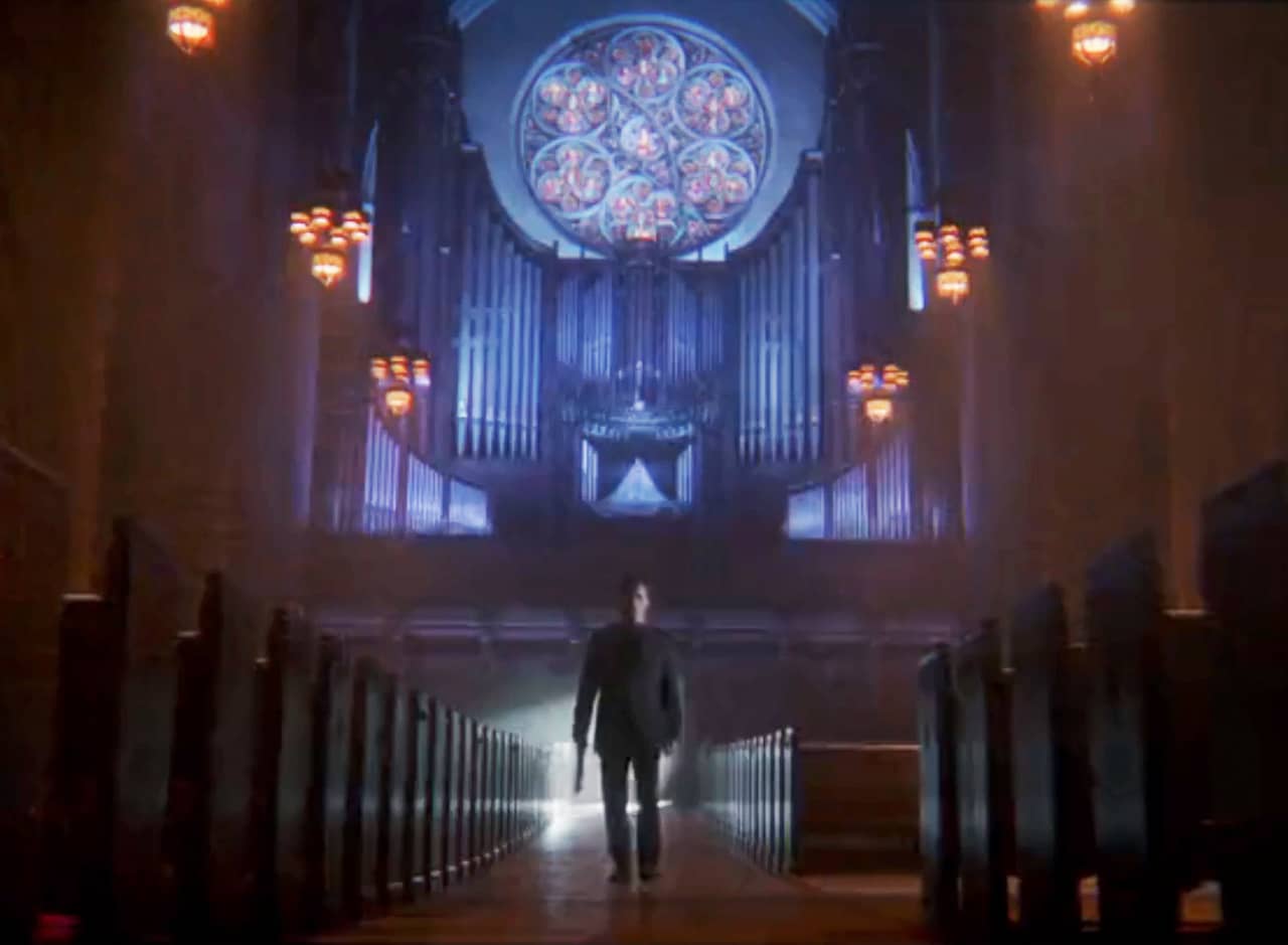 priest holding a gun walks down the aisle of a dark church with big stained glass window behind