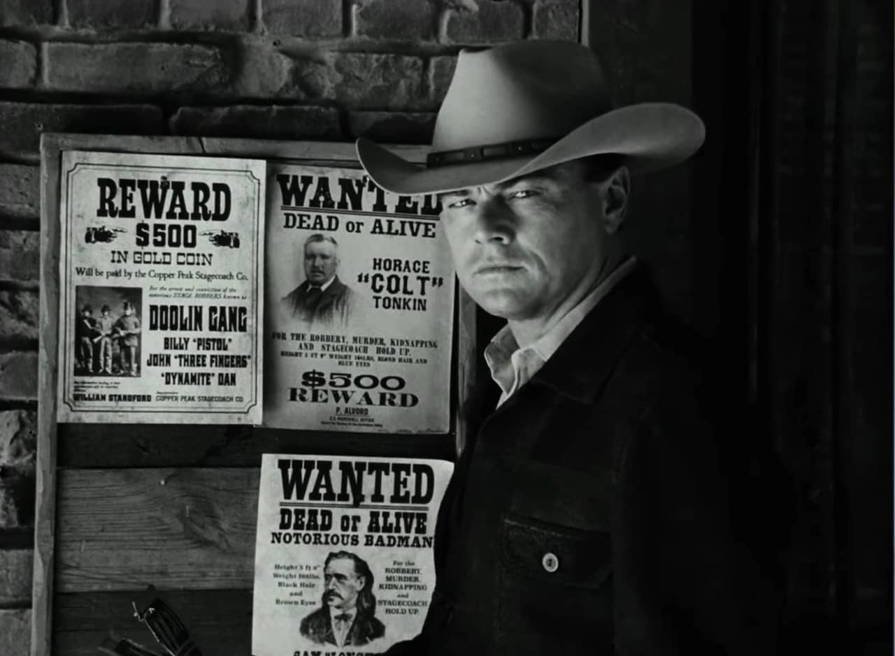 Rick Dalton as Jake Cahill in a cowboy hat stands in front of a bulletin board with Wanted posters