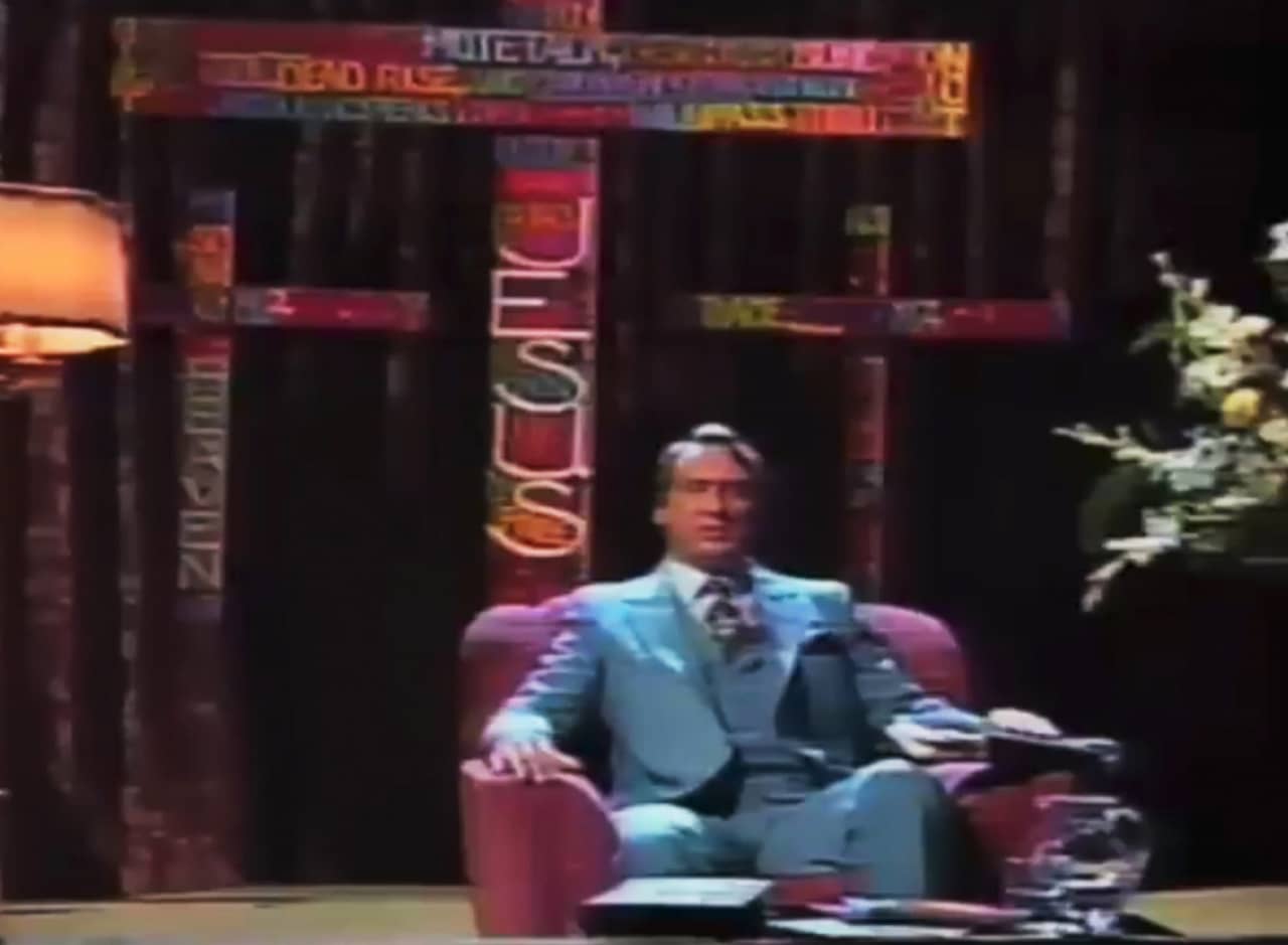 Brother Jim in a blues suit and sitting in an armchair on set in front of three colorful crosses