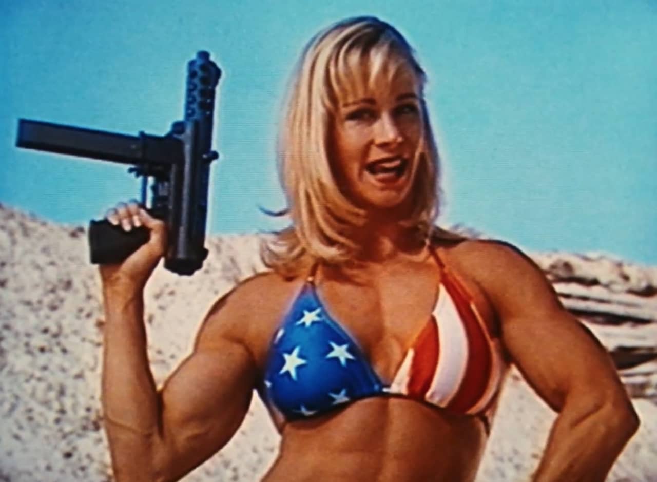 a blonde, muscular woman in an American flag bikini holds up a TEC-9 gun