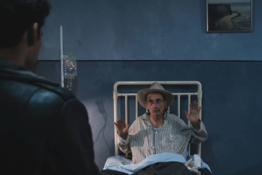 beat-up man in a hospital bed raising his hands during an interview
