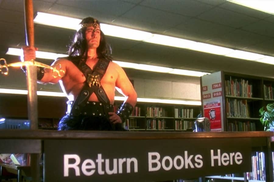 Conan poses with his sword in front of a “Return books here” sign