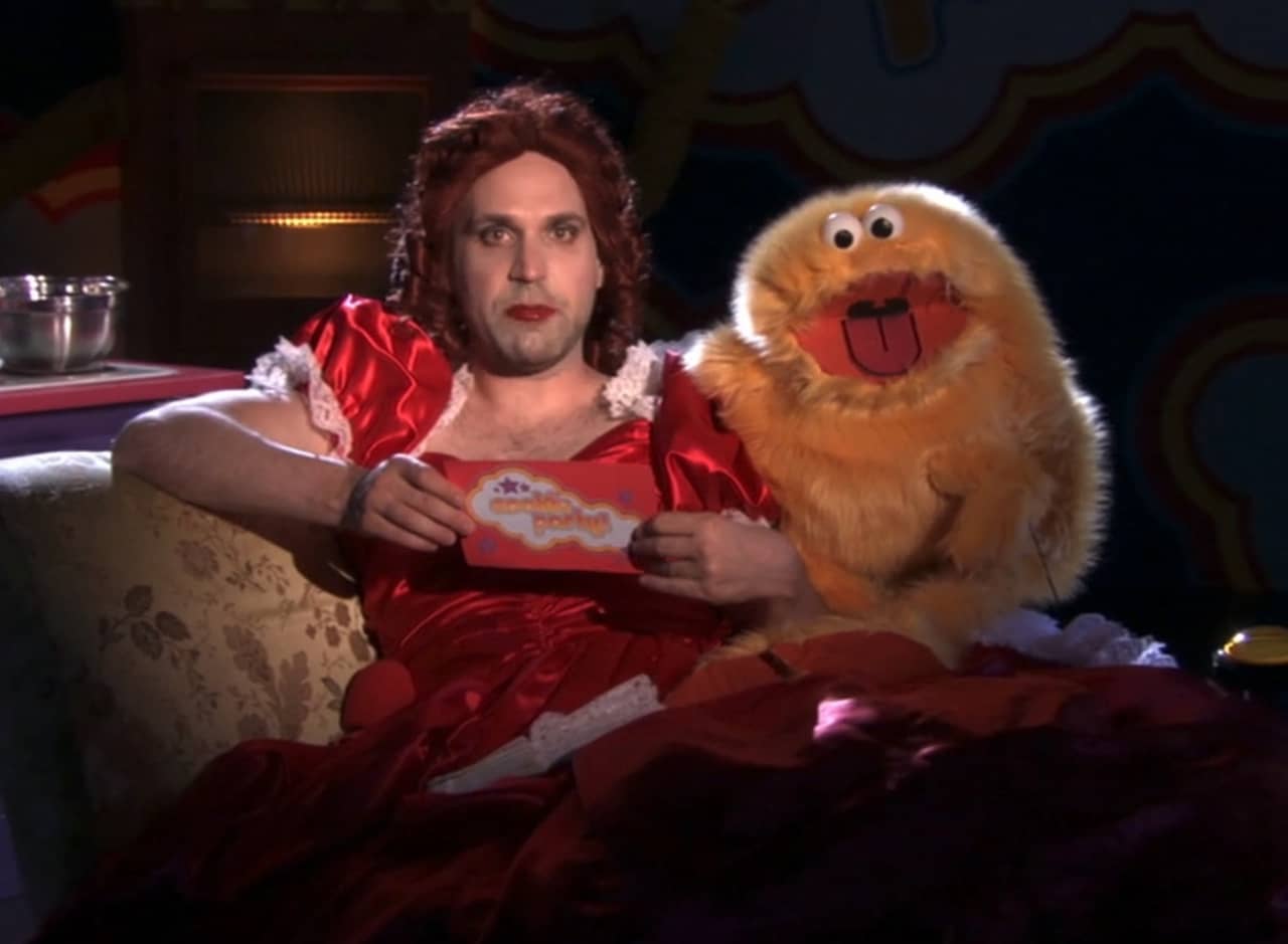 drag queen, Mini Coffee, in a satin red dress next to Ookie, an orange furry puppet