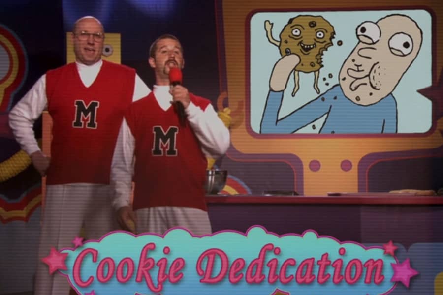 two men in matching sweater vests hold a mic and give their cookie dedication