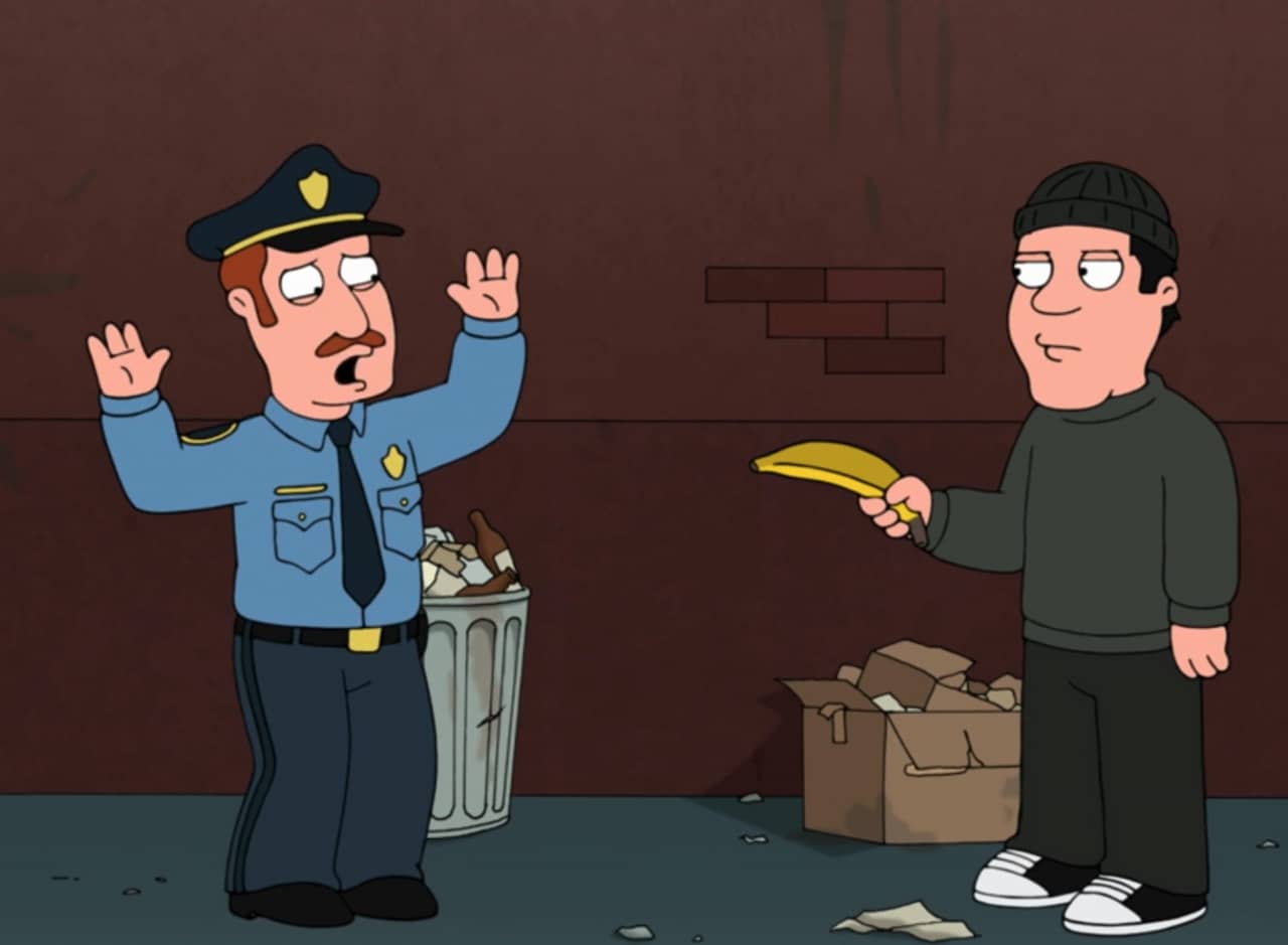 a man in a beanie points a banana at a scared cop with arms raised
