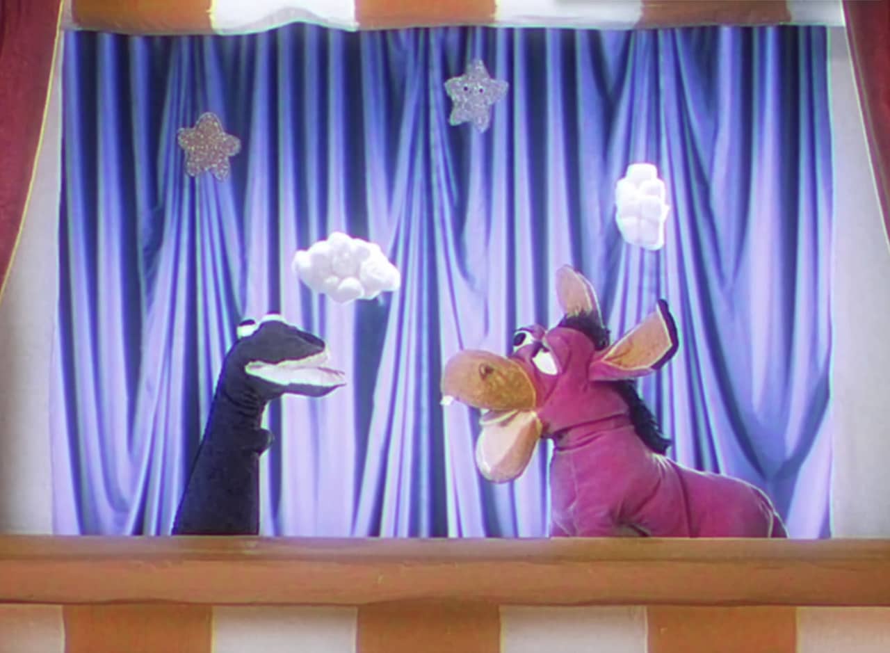 a puppet show with a pink donkey puppet and a black snake puppet in front of a blue curtain and cotton ball clouds