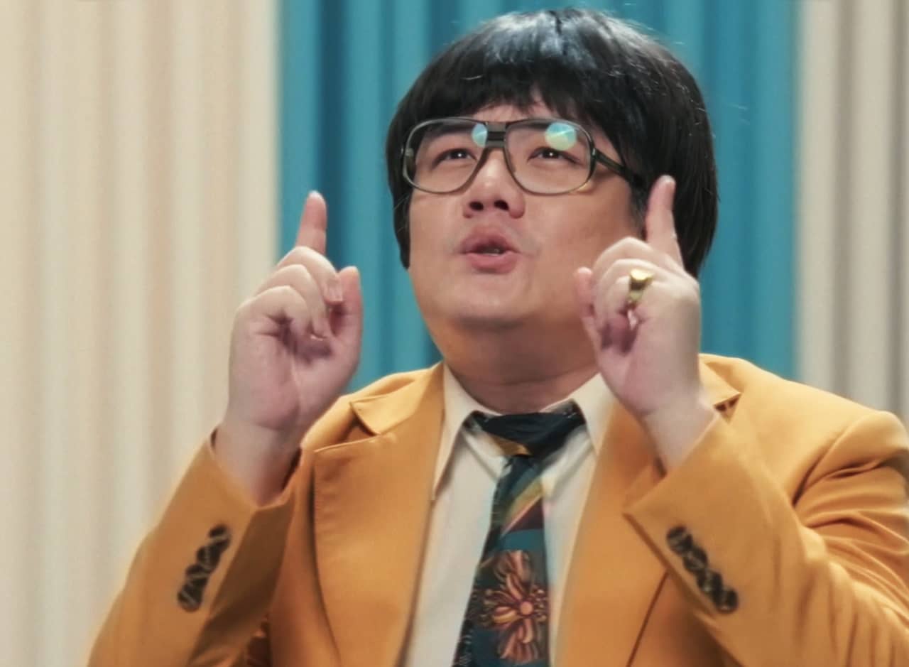 host who is an Asian man in a bright ’70s yellow suit and black-rimmed glasses, with a bowl cut