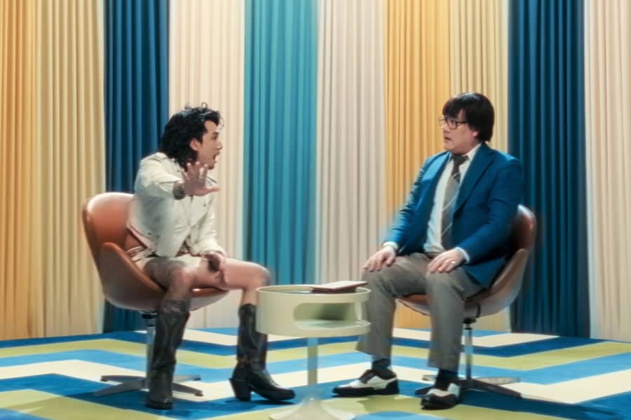 host with a musical guest: a young man with long hair in short shorts and tall boots