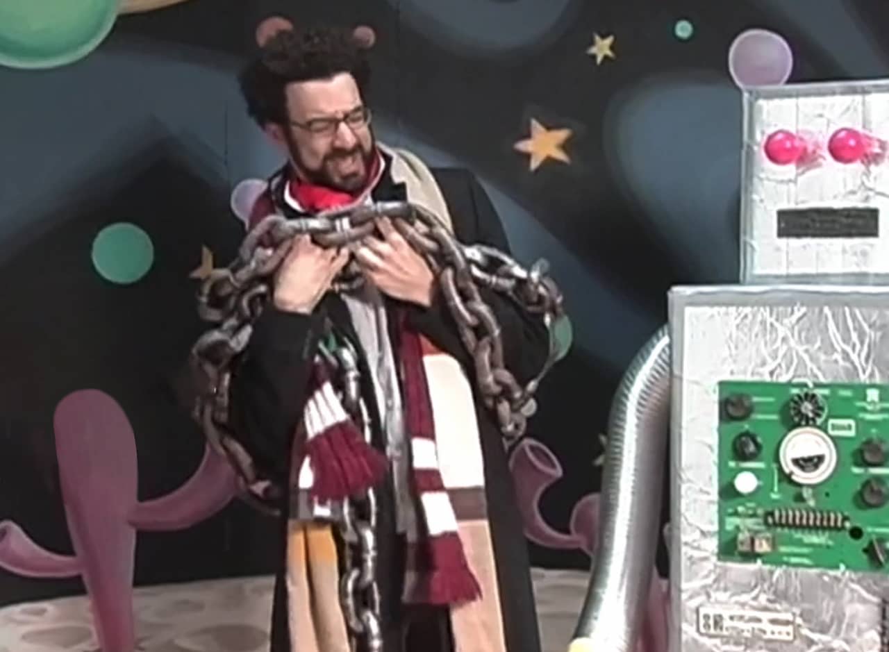 The Doctor, in a coat and scarf, struggling with big chains next to a robot