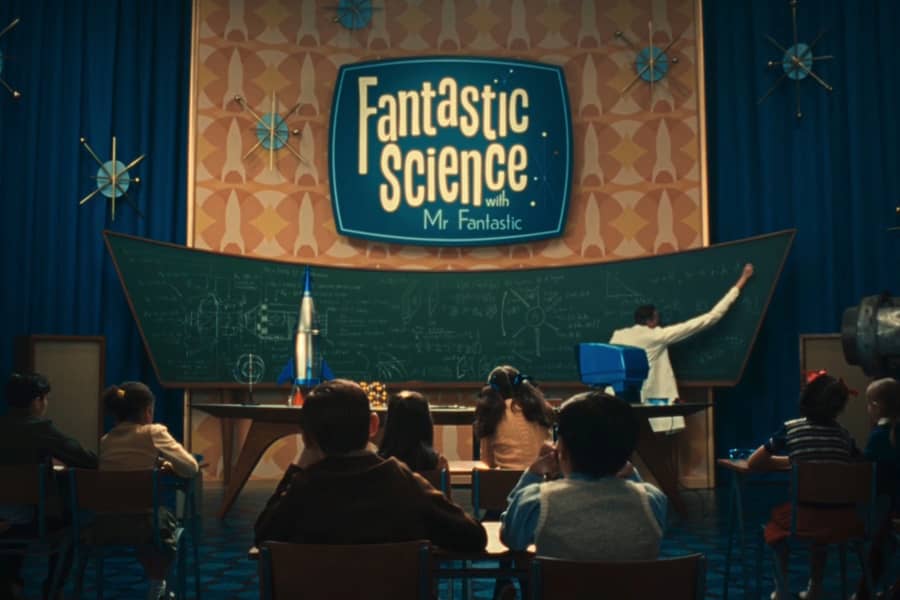the set of the show with kids in seats, Richards stretches his arm to reach a corner of the blackboard