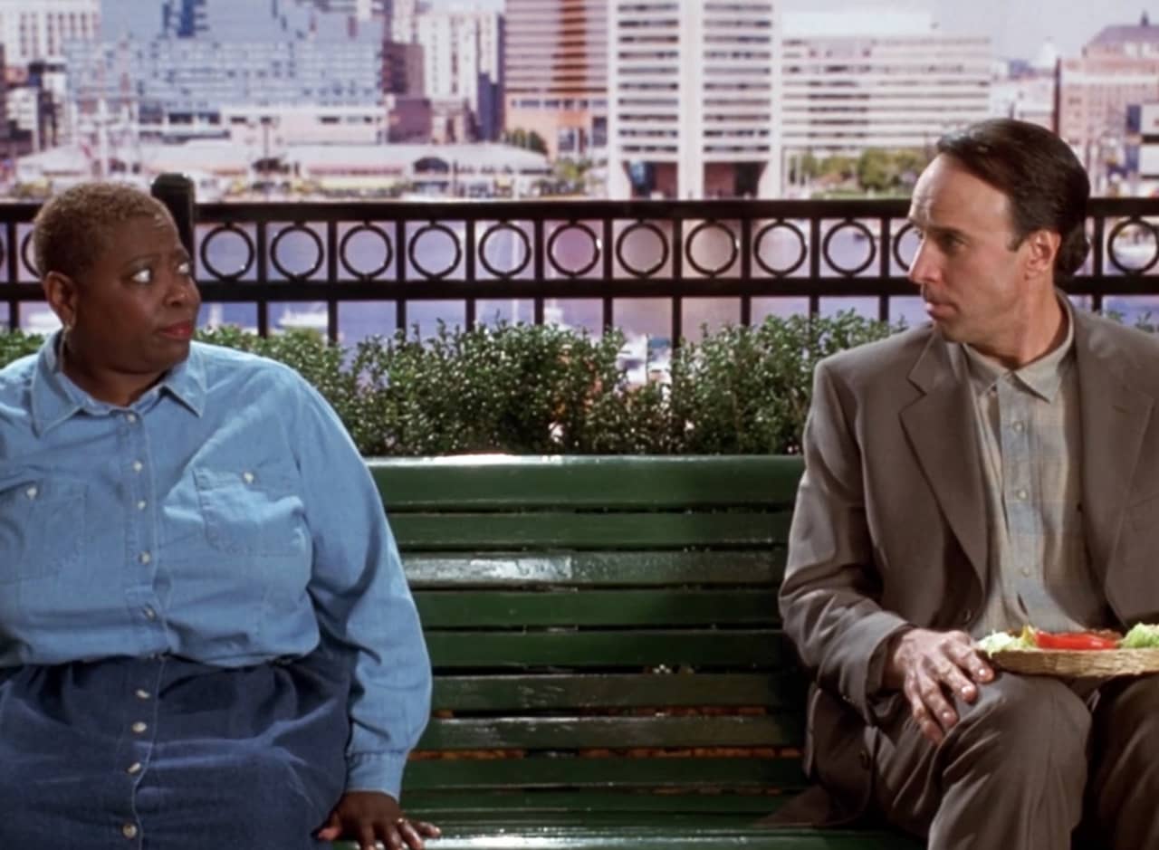 Kevin Nealon as Forrest Gump sitting on a bench and talking to a woman 