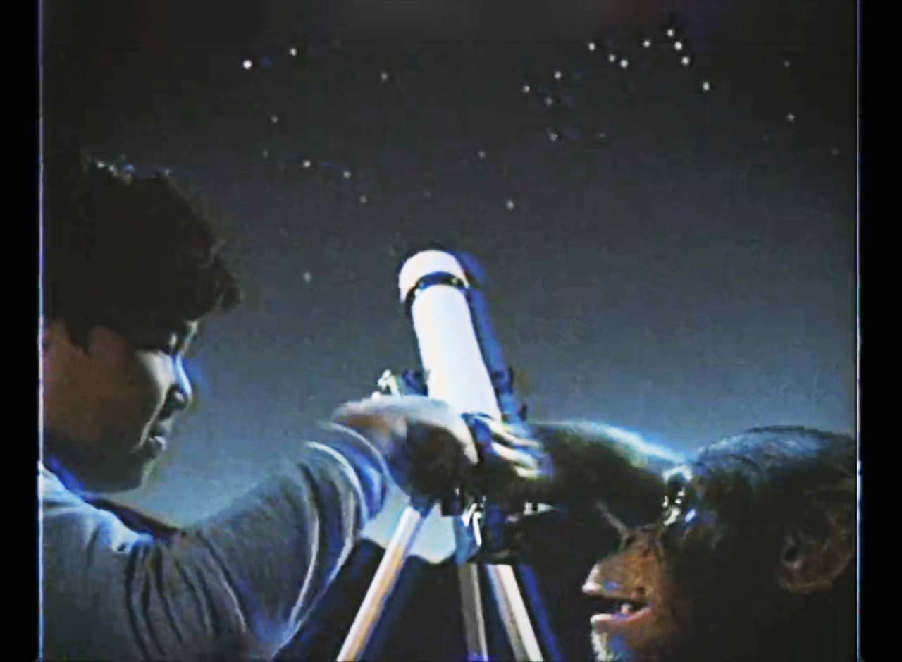 an Asian boy and a chimp fist bump in front of a telescope and night’s sky