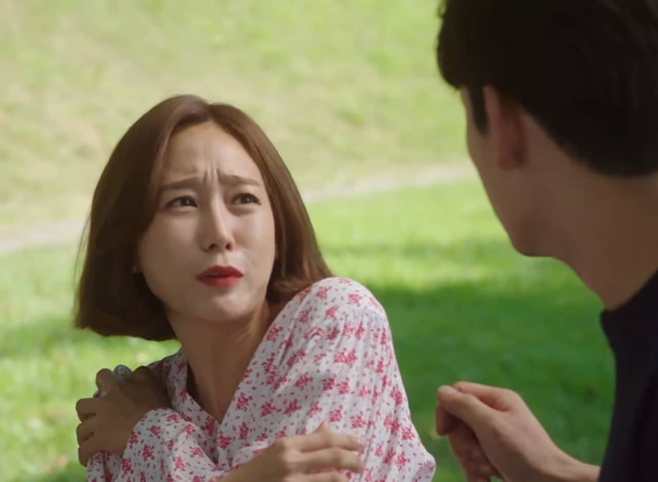 a Korean man and woman sit in the park and the woman recoils in pain