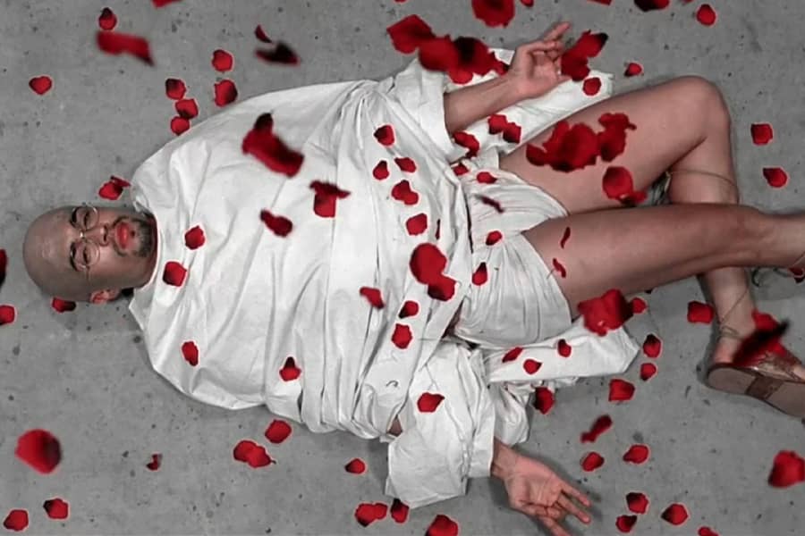 Gandhi lays on the ground in his robes as red flower petals fall on him romantically