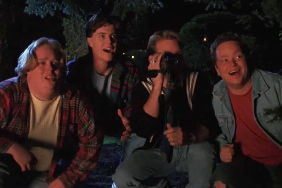 four college dudes crouching outside at night, one is holding a video camera
