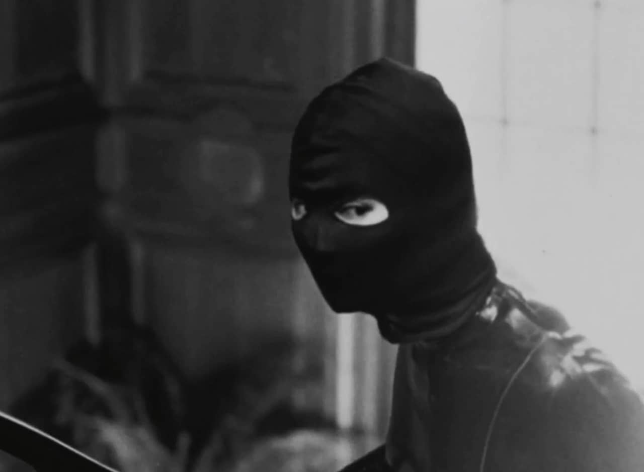 black and white film: a woman in a leather catsuit and a ski mask walks up a flight of stairs