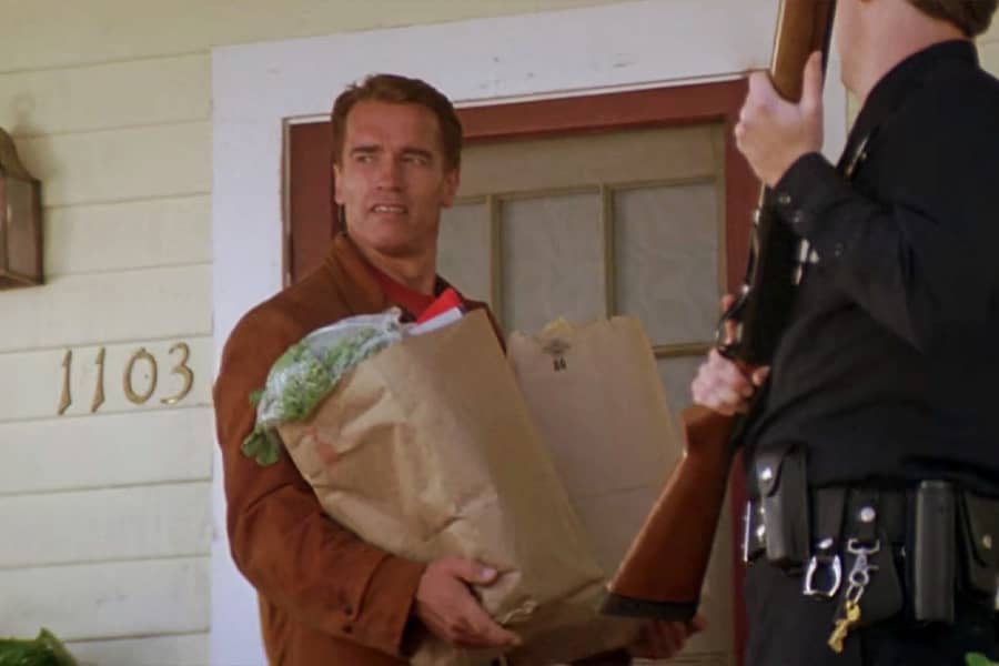 Slater holding bags of groceries on a porch, a cop with a gun stands by