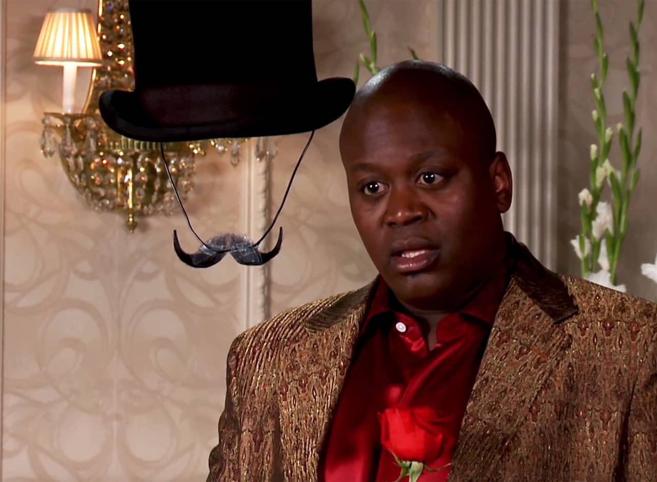 contestant Titus Andromedon talking with Invisible Joe, who can only be seen by his top hat and mustache that is taped to the chin strap of the hat