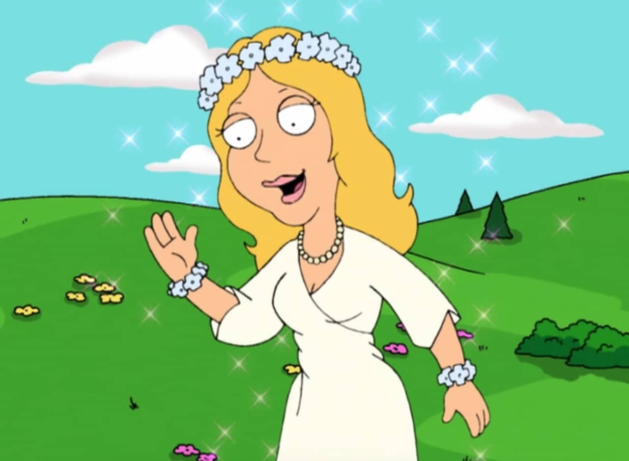 Mother Maggie, a sparkling woman who wears all white and a flower crown in front of rolling hills of green grass
