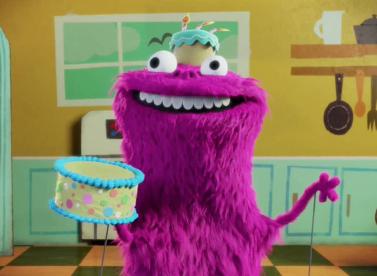 Kakie, a pink furry monster puppet with a cake hat, smiles and holds a birthday cake in a fake kitchen