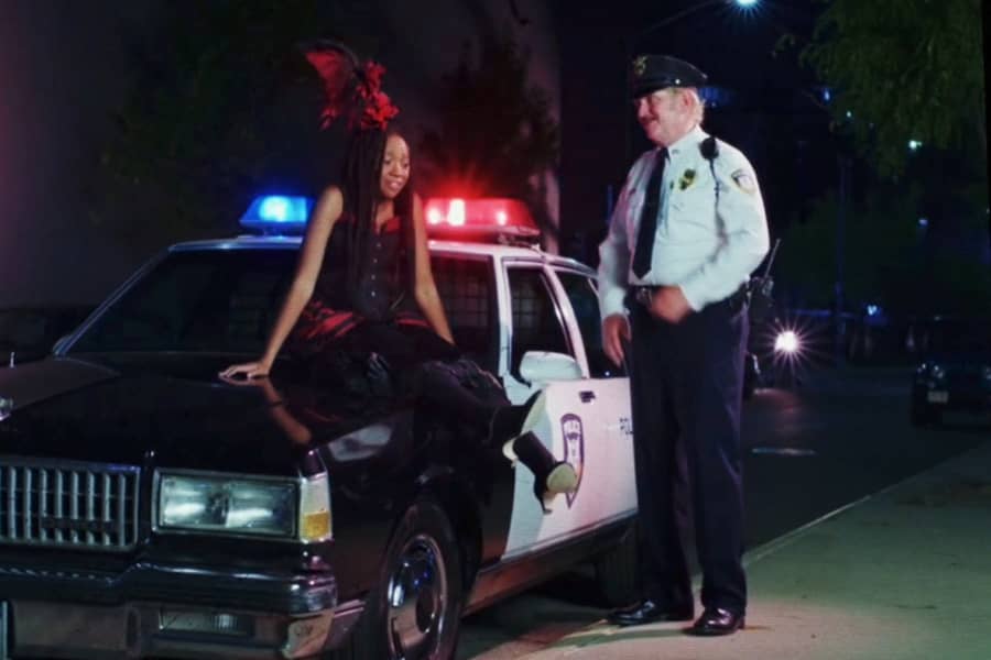 Cassidy dressed in French dancer costume, sitting on the hood of a cop car with the chief