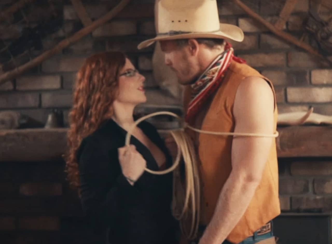 curly red-haired woman holding a lasso that’s wrapped around a cowboy man