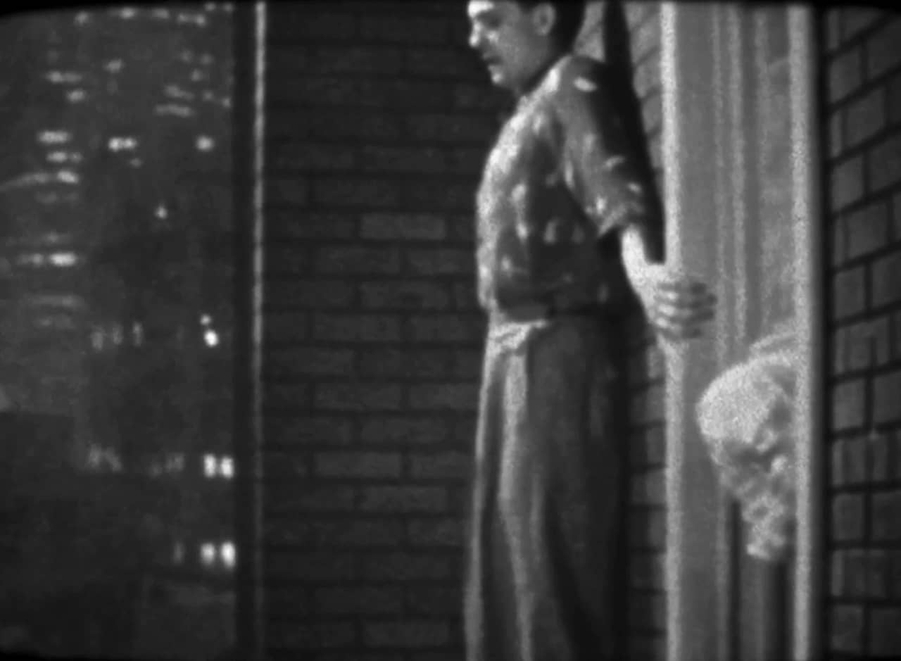 man outside a building, gripping the brick wall; a woman pokes her head out a nearby window