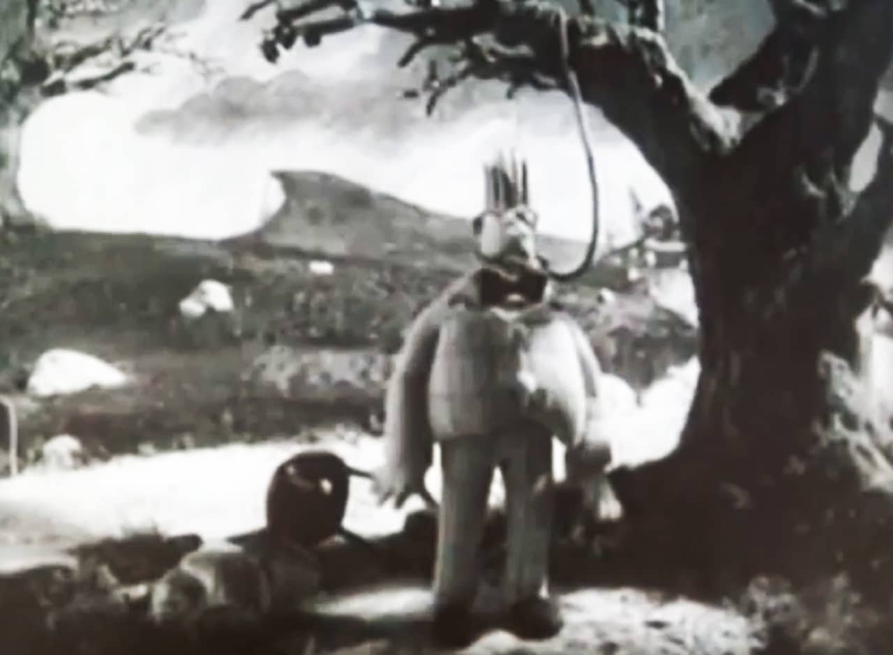 Arnold with a noose around his neck, tied to a tree branch, but the rope is too long so he is standing on the ground unharmed
