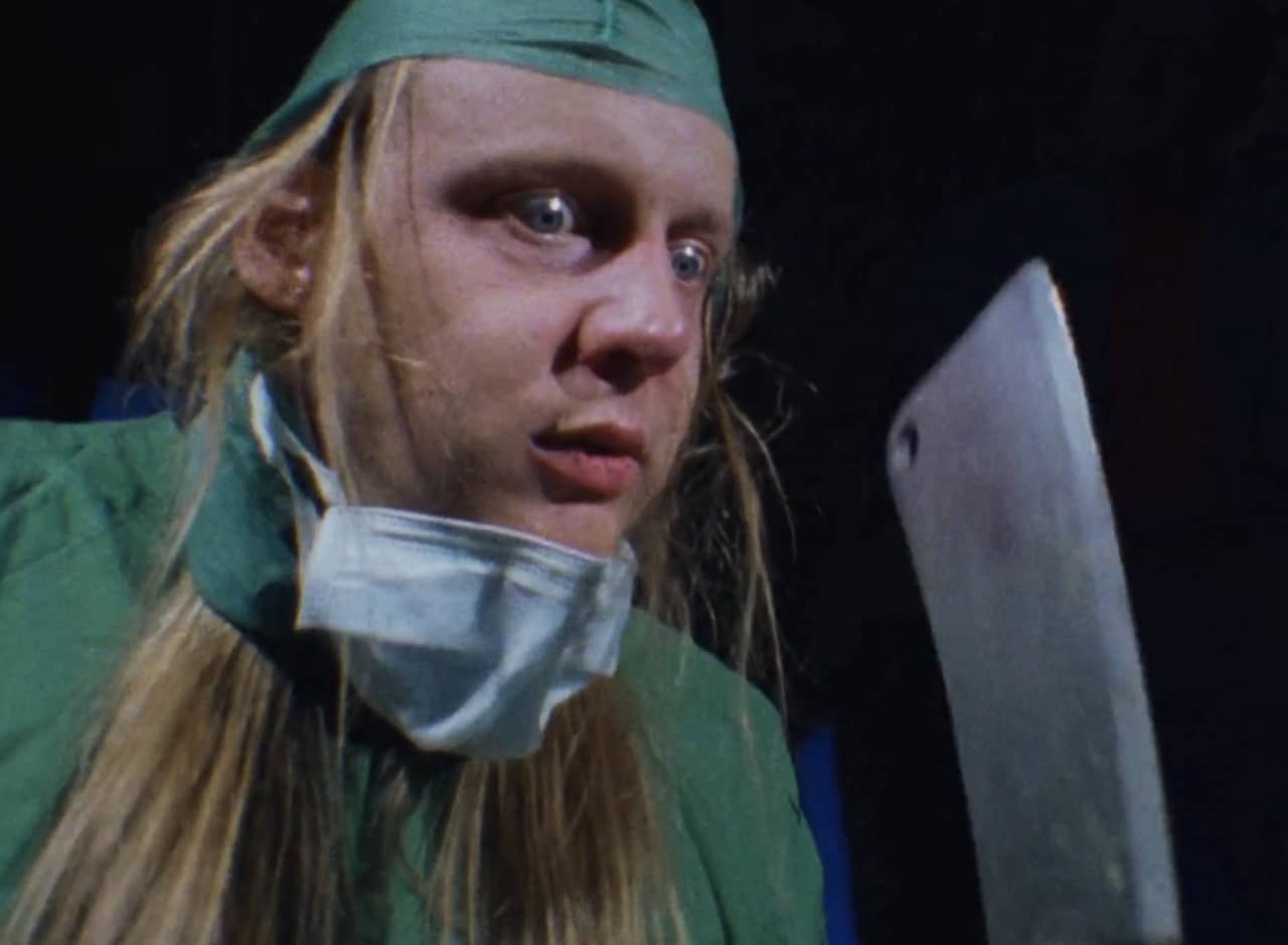 a long-haired, wide-eyed man in scrubs holding a cleaver