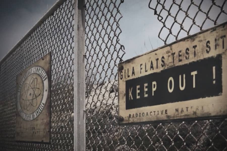 a chain link fence with a hole in it and a sign that reads “Gila Flats test site keep out