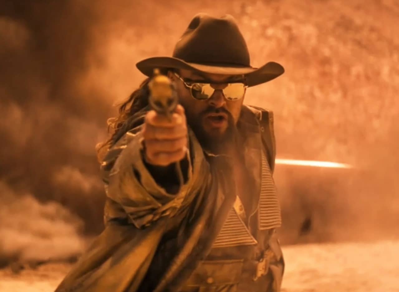 Jason Momoa, color-graded in orange, wearing sunglasses and a cowboy hat aiming a gun at the camera