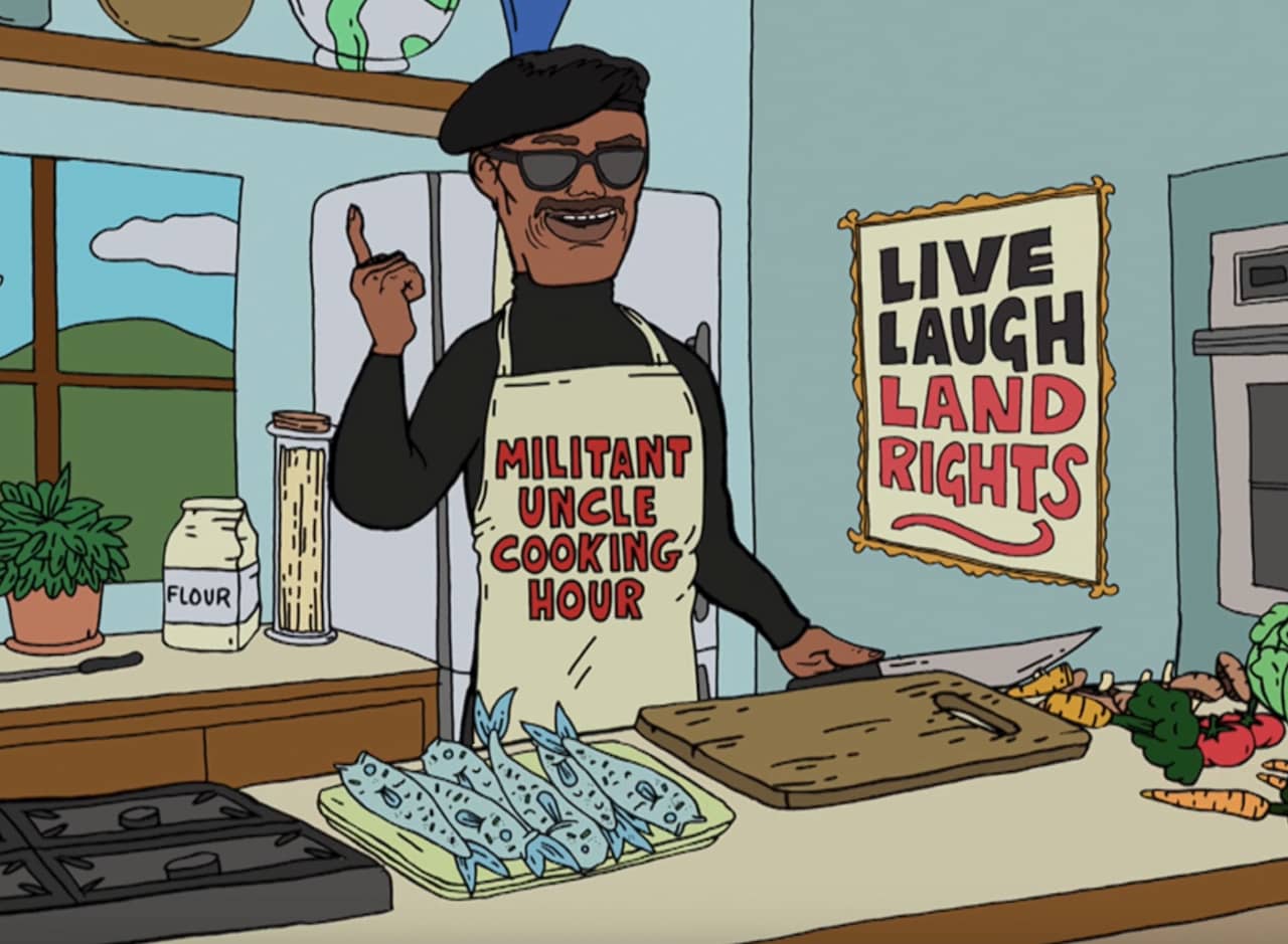 Uncle Noogy, a Māori man wearing a beret and sunglasses, preparing a dish in the kitchen; there is a “Live, Laugh, Land Rights” sign on the wall