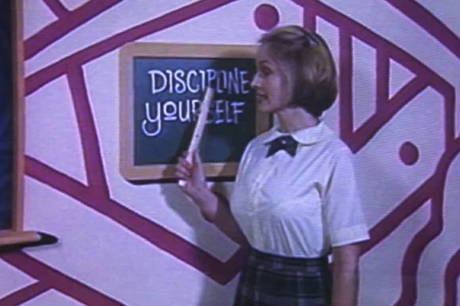 Miss Sally, in a schoolgirl outfit, points at a blackboard that says “Discipline yourself”