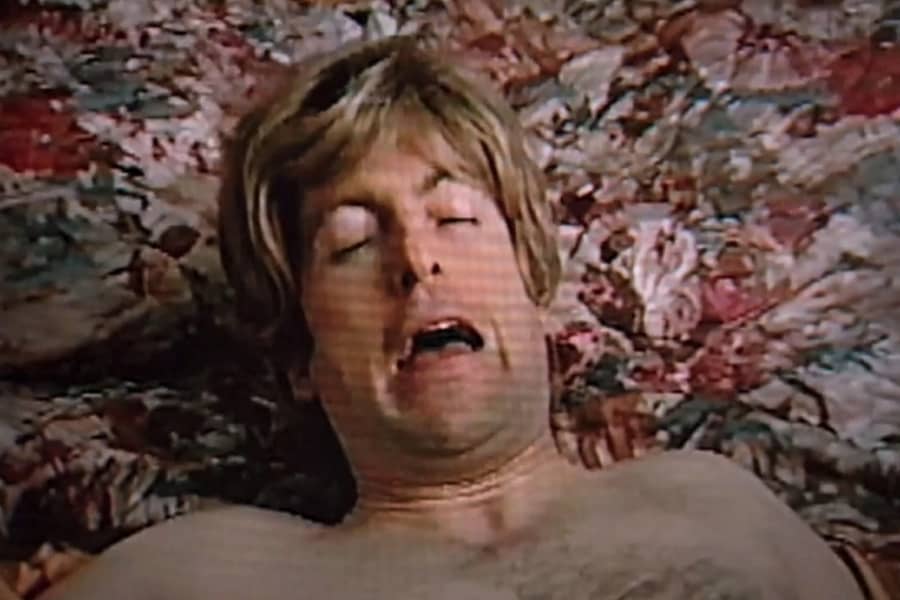 Gil lying shirtless on the bed, making a silly face