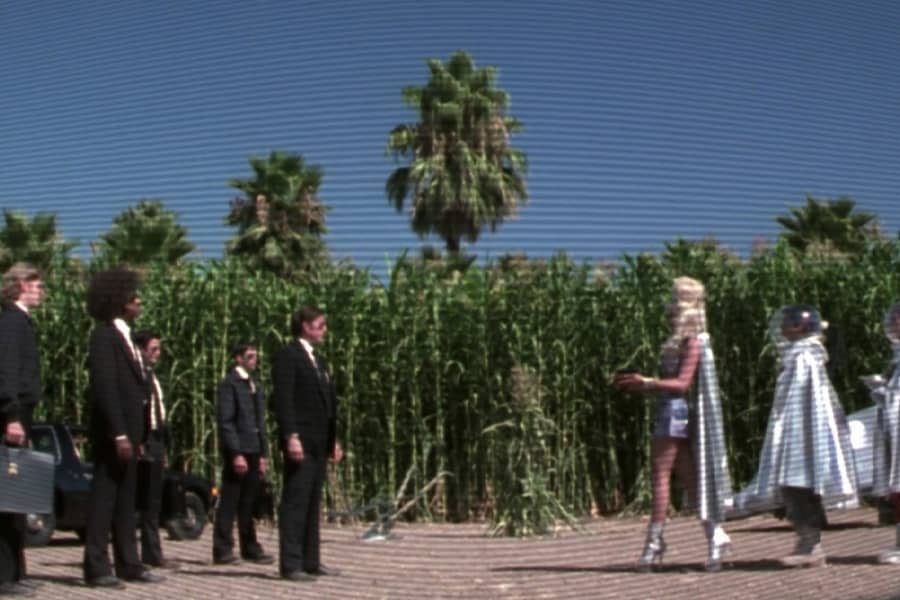 against a cornfield, a group of “aliens” in sliver clothing approach a group of men dressed in black suits and sunglasses