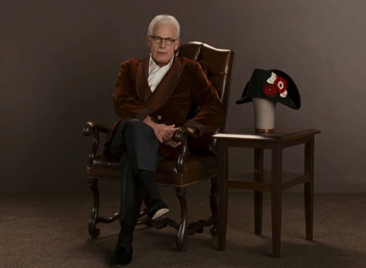 elderly man in a leather armchair and smoking jacket speaks at the camera